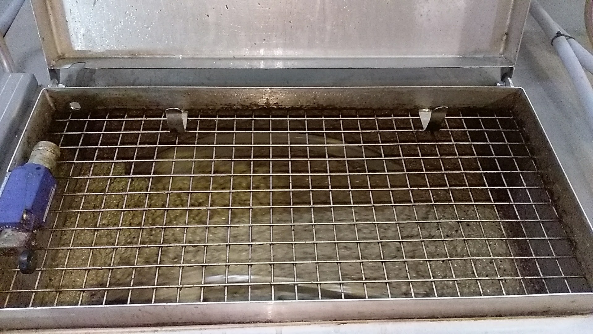 An open metal container with a grid top, containing dirty liquid and a blue valve attachment on the side.